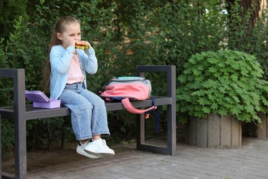 Photo of Cute little girl eating sandwich on bench outdoors. School lunch