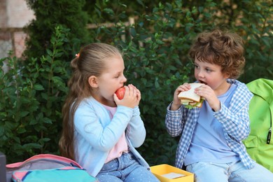 Photo of Cute kids eating food on bench outdoors. School lunch