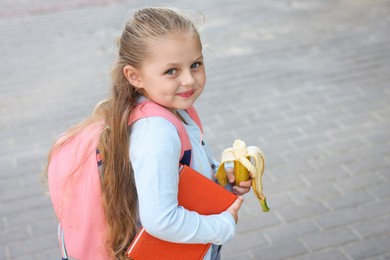Photo of Cute little girl eating banana outdoors, space for text. School lunch