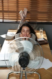 Photo of Woman enjoying air flow from fan while lying on table at workplace. Summer heat