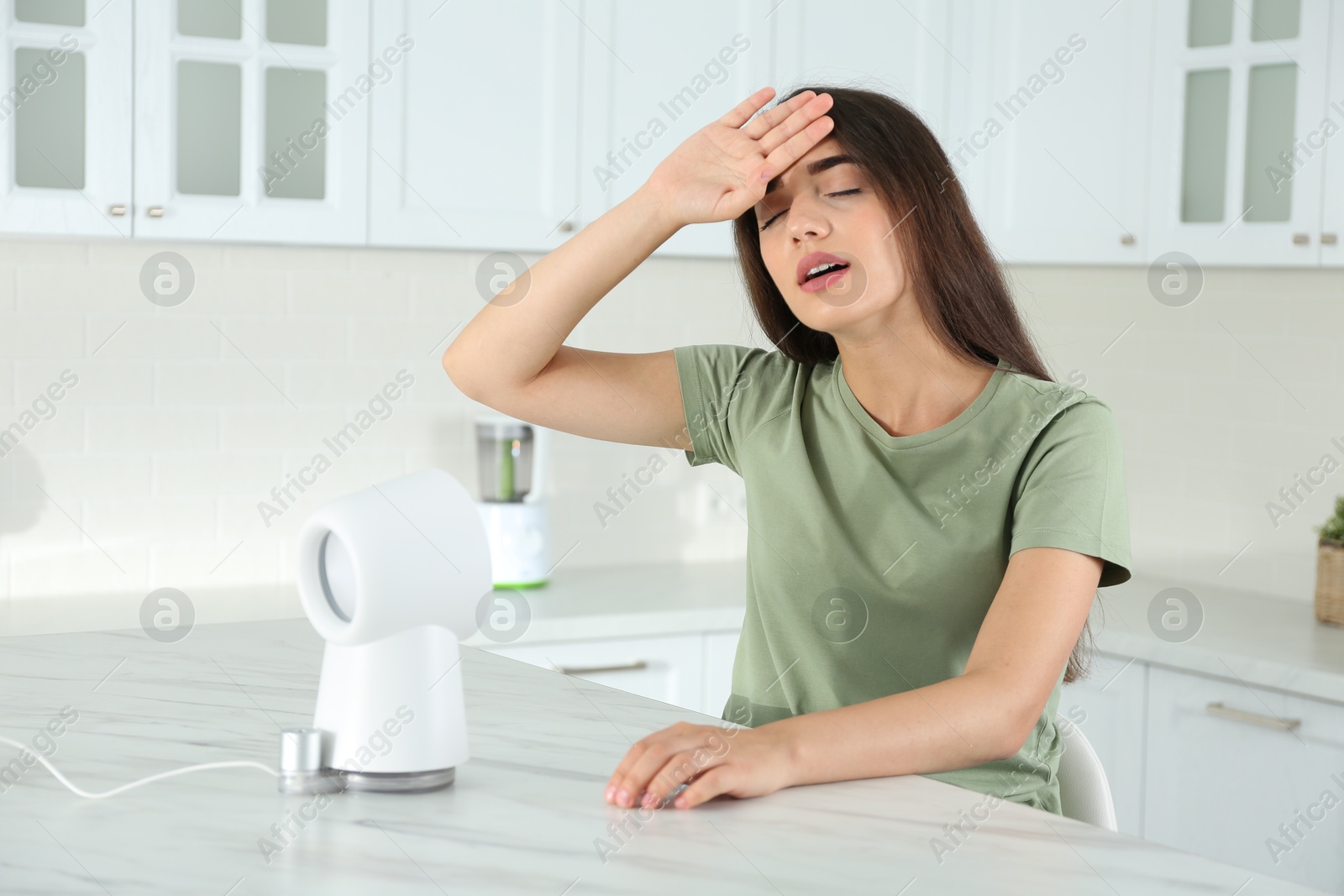Photo of Woman with portable fan suffering from heat in kitchen. Summer season