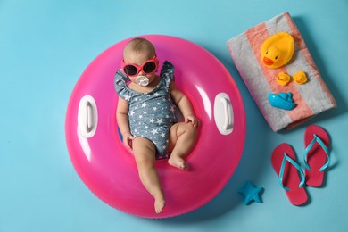Photo of Cute little baby in sunglasses with inflatable ring and beach accessories on light blue background, top view