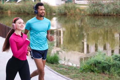 Photo of Smiling couple in headphones running near river outdoors. Space for text