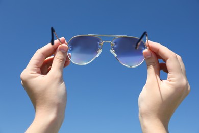 Photo of Woman holding stylish sunglasses against blue sky, closeup
