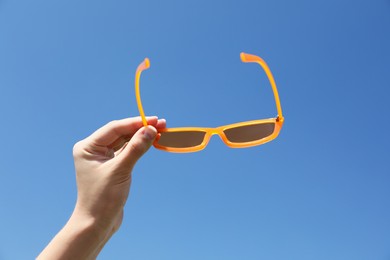 Photo of Woman holding stylish sunglasses against blue sky, closeup