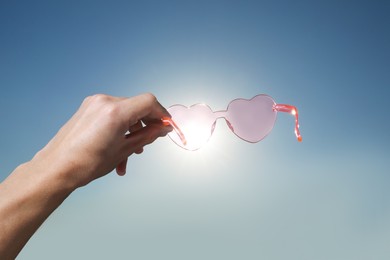 Photo of Woman holding stylish heart-shaped sunglasses against blue sky, closeup