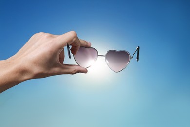 Photo of Woman holding stylish heart-shaped sunglasses against blue sky, closeup
