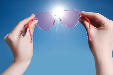 Photo of Woman holding stylish heart-shaped sunglasses against blue sky, closeup