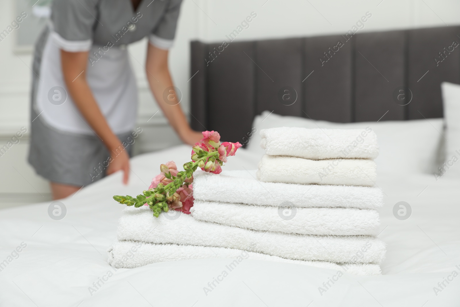 Photo of Chambermaid working in hotel room, focus on stack of terry towels and beautiful flowers on bed