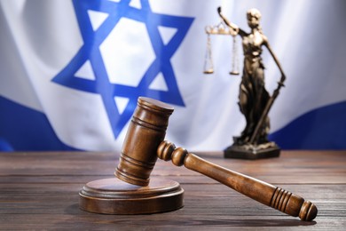 Photo of Judge's gavel and statue of Lady Justice on wooden table against flag of Israel, closeup