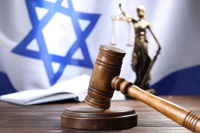 Photo of Judge's gavel, book and statue of Lady Justice on wooden table against flag of Israel, closeup