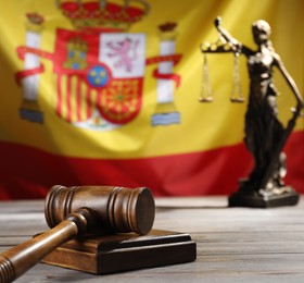 Photo of Judge's gavel and statue of Lady Justice on wooden table against flag of Spain, closeup