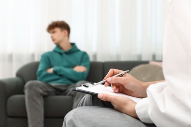 Photo of Professional psychologist working with teenage boy and writing in clipboard in office, selective focus