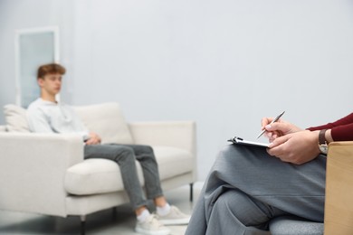 Photo of Professional psychologist working with teenage boy and writing in clipboard in office, selective focus