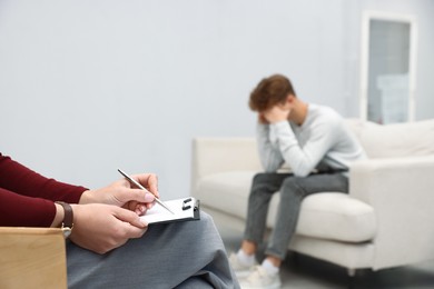 Photo of Professional psychologist working with teenage boy and writing in clipboard in office, selective focus