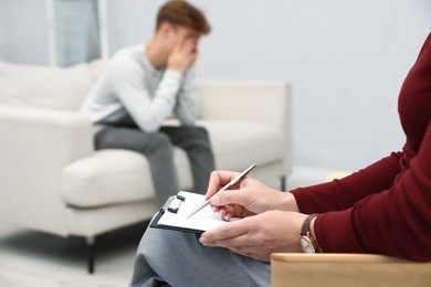 Photo of Professional psychologist working with teenage boy and writing in clipboard in office, selective focus