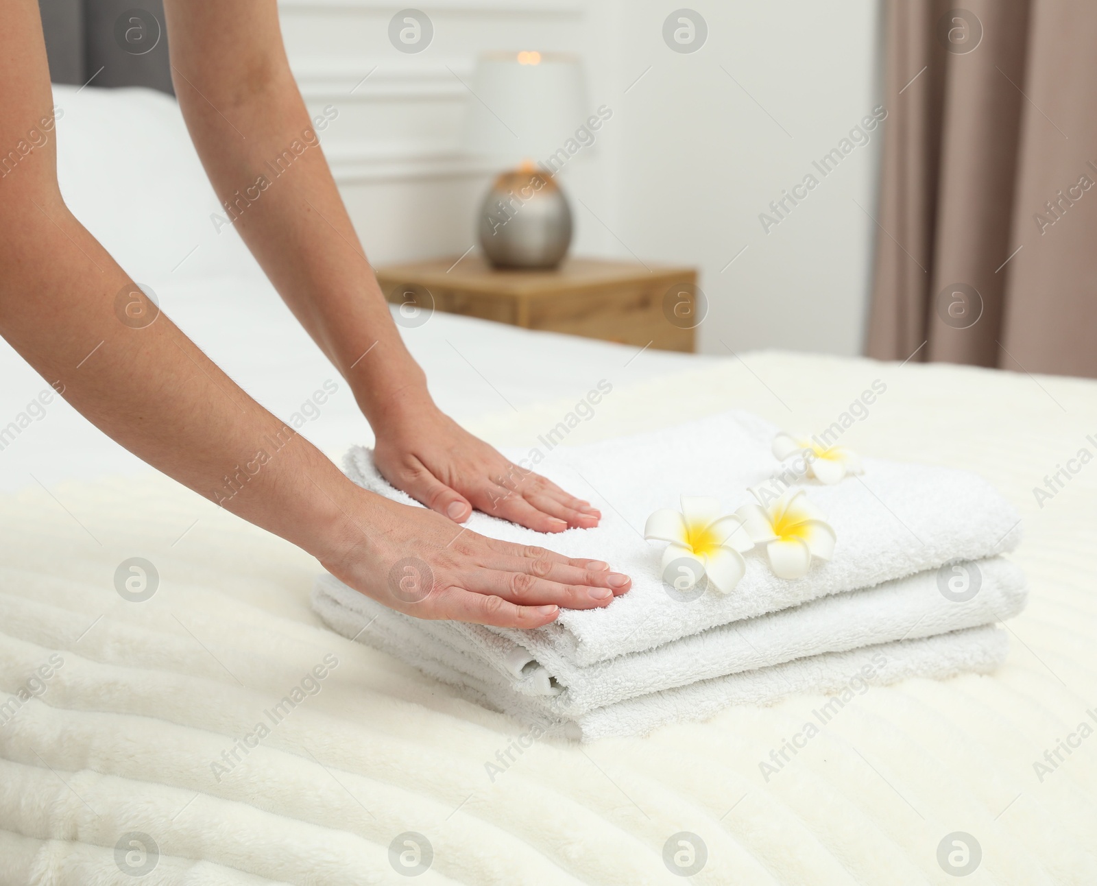 Photo of Woman putting stack of white terry towels and plumeria flowers onto bed indoors, closeup