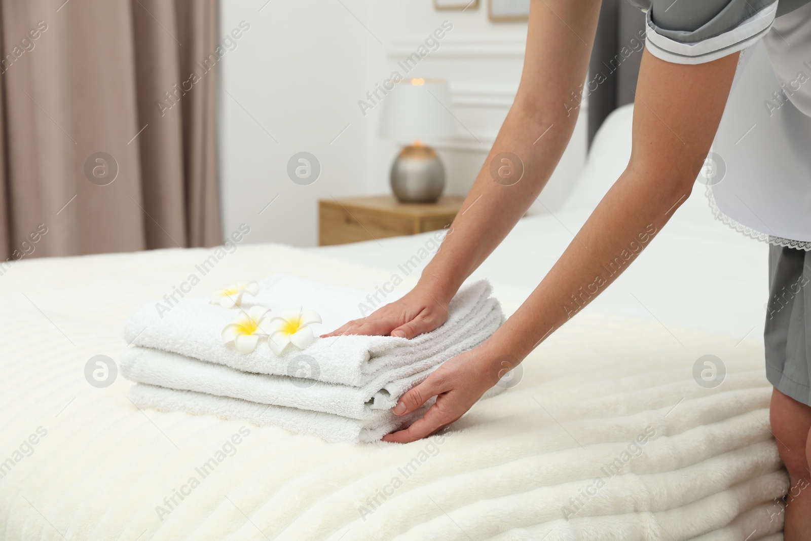Photo of Chambermaid putting stack of white terry towels and plumeria flowers onto bed indoors, closeup