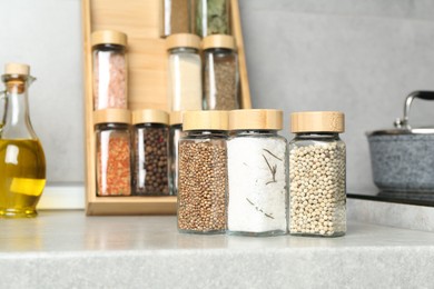 Photo of Different spices in glass jars and oil on light grey table in kitchen, closeup