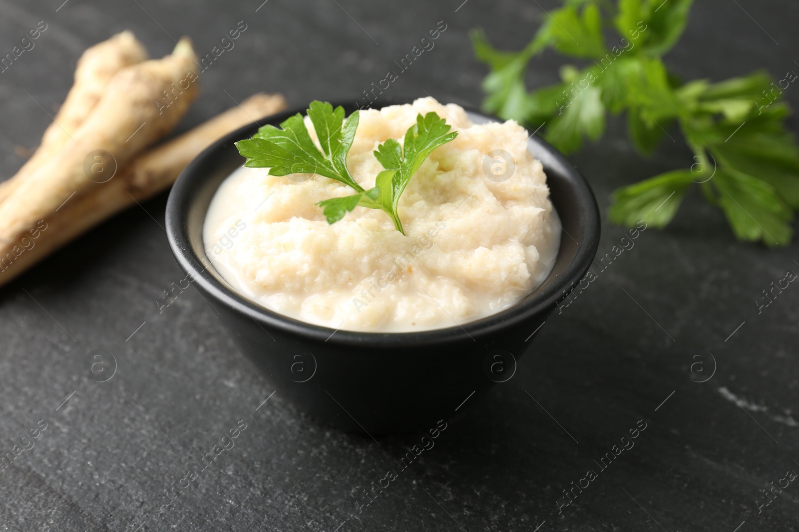 Photo of Tasty horseradish sauce with parsley and roots on black table, closeup