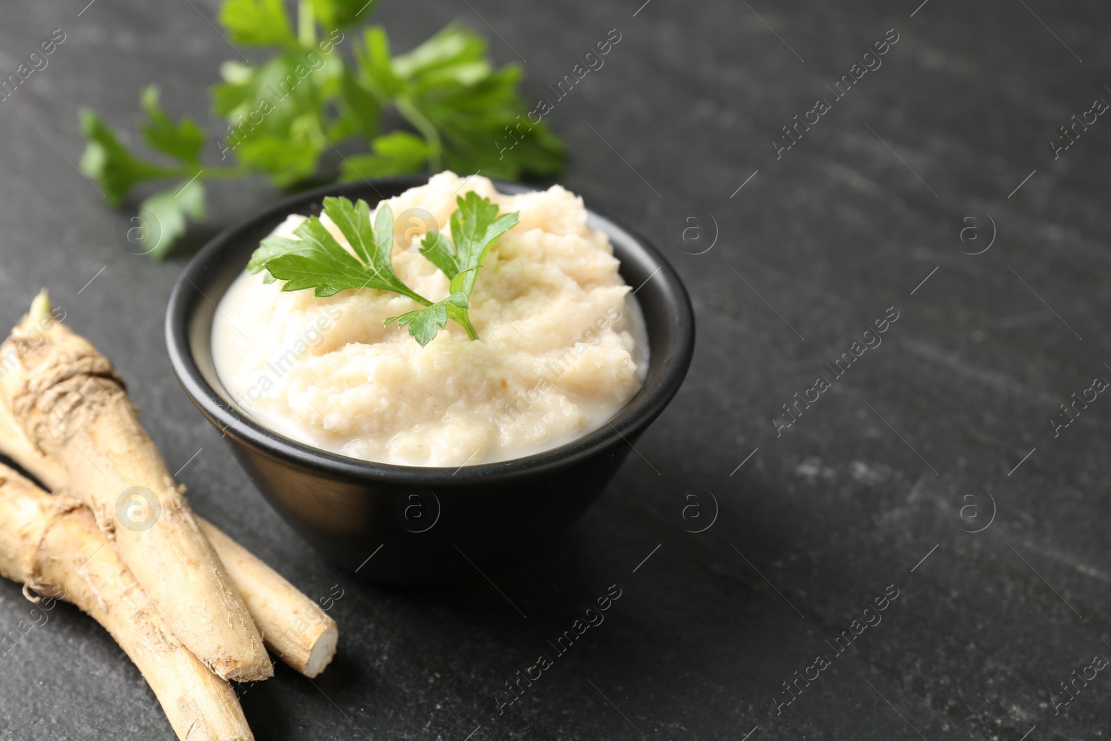 Photo of Tasty horseradish sauce with parsley and roots on black table, closeup. Space for text