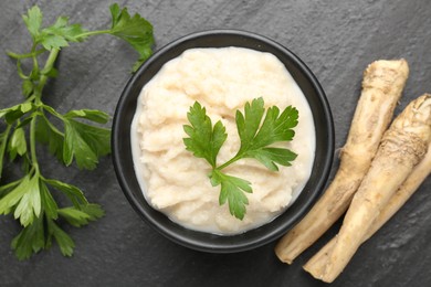 Photo of Tasty horseradish sauce with parsley and roots on black table, flat lay