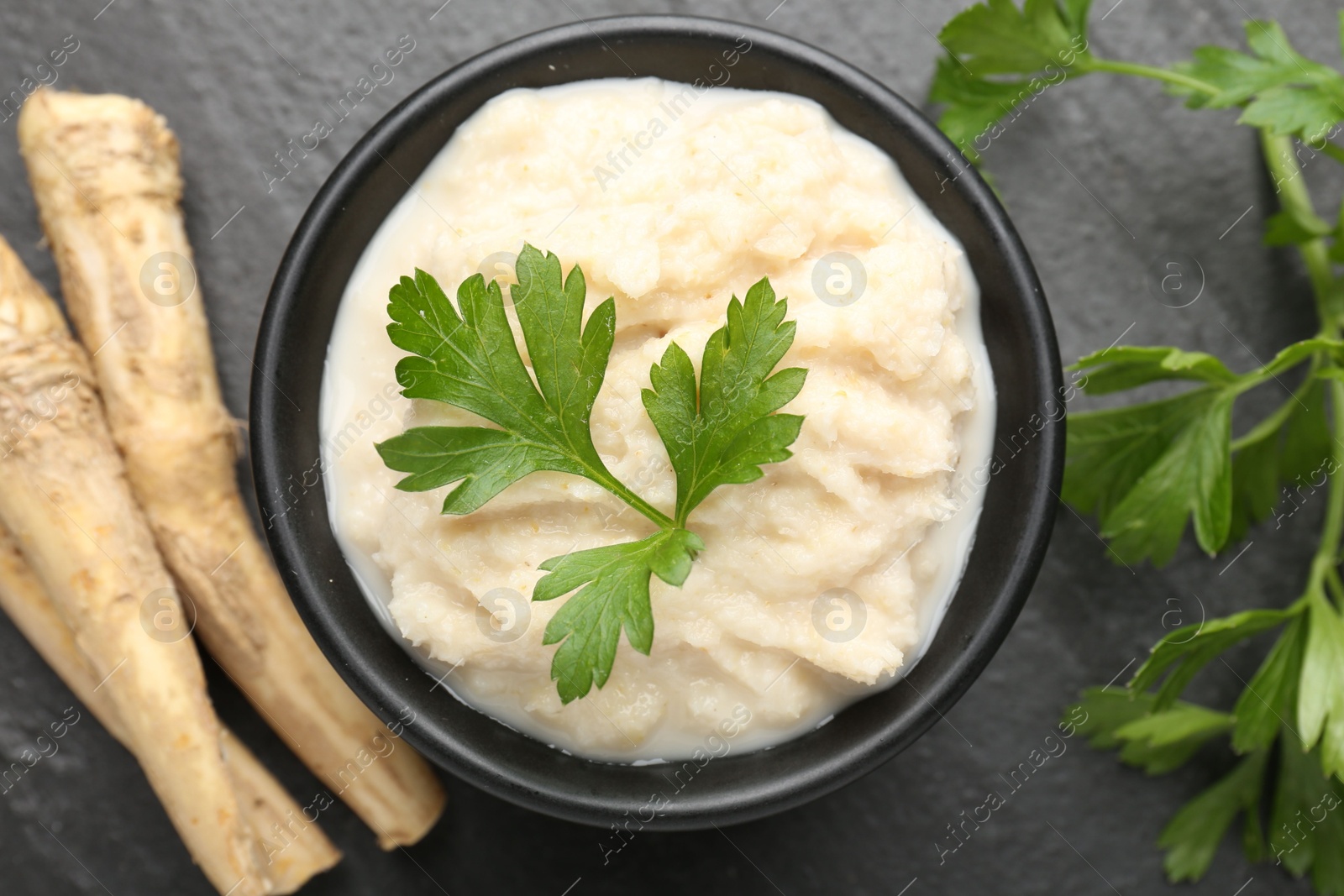 Photo of Tasty horseradish sauce with parsley and roots on black table, flat lay