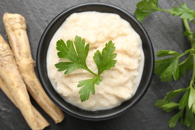 Photo of Tasty horseradish sauce with parsley and roots on black table, flat lay