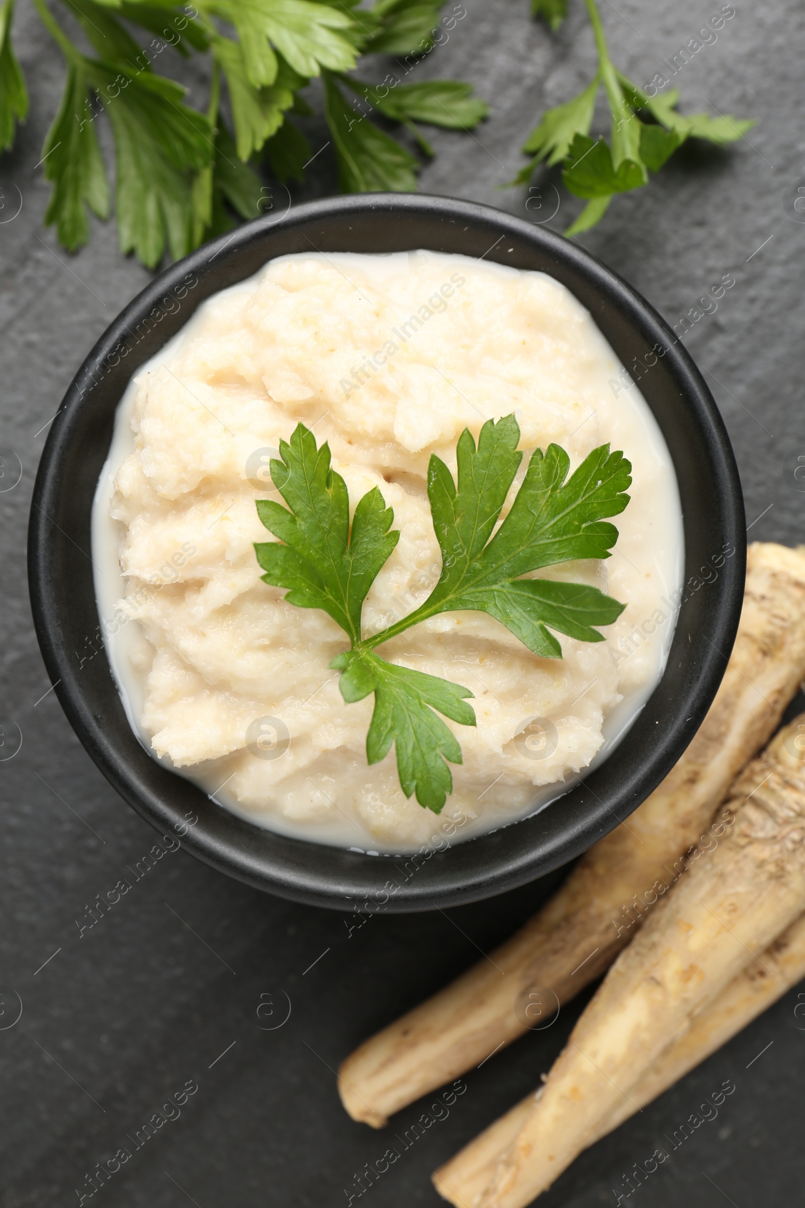 Photo of Tasty horseradish sauce with parsley and roots on black table, flat lay