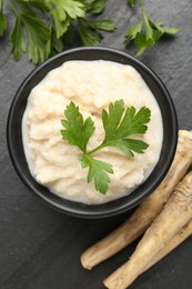Photo of Tasty horseradish sauce with parsley and roots on black table, flat lay