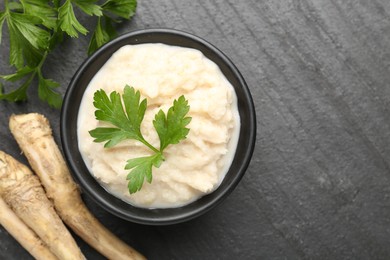 Photo of Tasty horseradish sauce with parsley and roots on black table, flat lay. Space for text