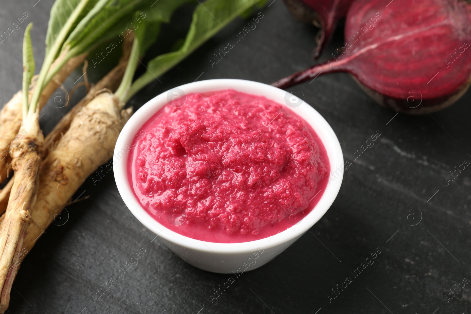 Photo of Tasty horseradish sauce with beetroot, roots and vegetable on black table, closeup
