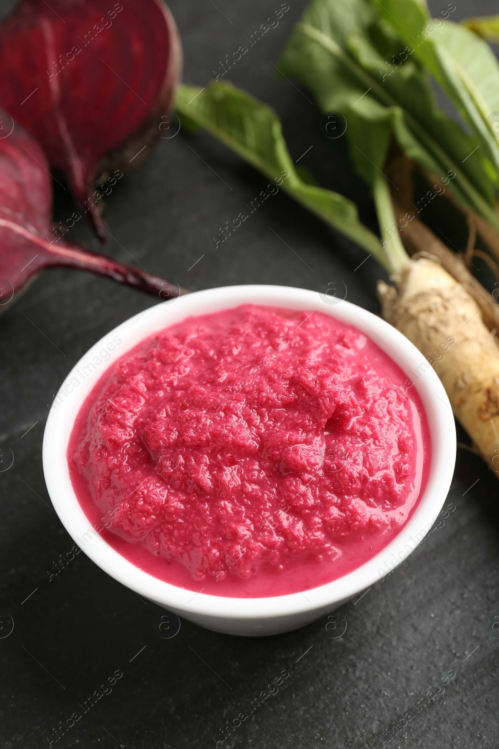 Photo of Tasty horseradish sauce with beetroot, roots and vegetable on black table, closeup