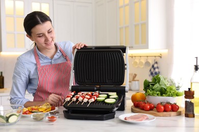 Photo of Woman cooking skewers with meat and vegetables on electric grill at home