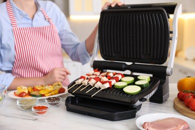 Photo of Woman cooking skewers with meat and vegetables on electric grill at home, closeup
