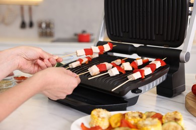 Photo of Woman cooking skewers with meat and vegetables on electric grill at home, closeup