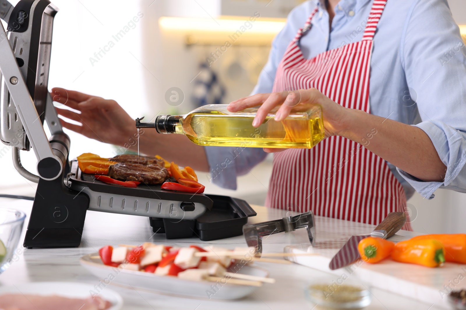 Photo of Woman pouring oil onto vegetables cooking on electric grill in kitchen, closeup