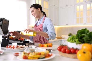 Photo of Woman pouring oil onto vegetables cooking on electric grill in kitchen