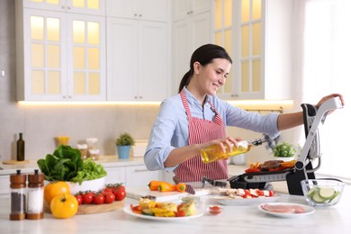 Photo of Woman pouring oil onto vegetables cooking on electric grill in kitchen