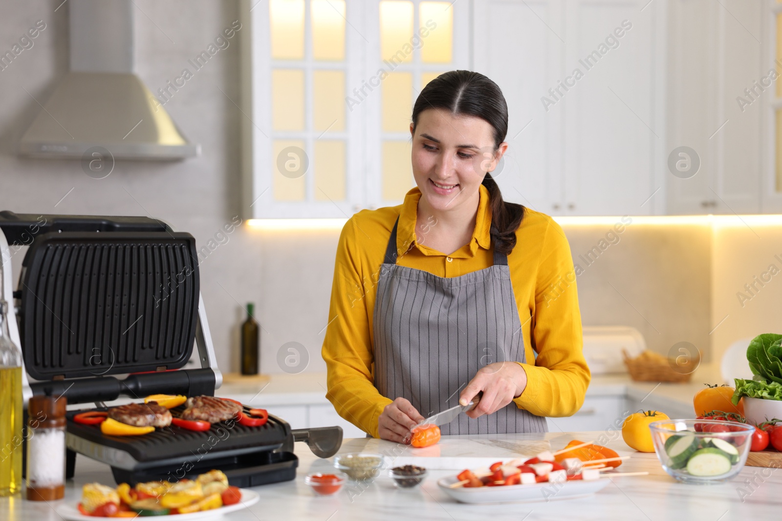 Photo of Woman cutting pepper while cooking meat and vegetables on electric grill in kitchen