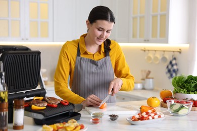 Photo of Woman cutting pepper while cooking meat and vegetables on electric grill in kitchen