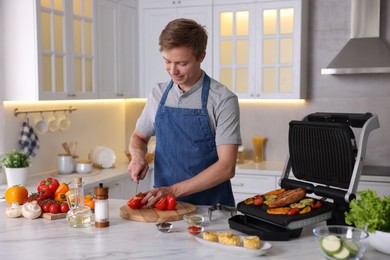 Photo of Man cutting pepper while cooking meat and vegetables on electric grill in kitchen
