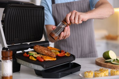 Photo of Man adding pepper to vegetables and meat cooking on electric grill in kitchen, closeup