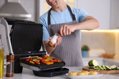 Photo of Man adding salt to vegetables cooking on electric grill in kitchen, closeup