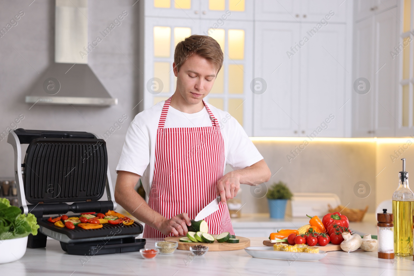 Photo of Man cutting zucchini while cooking vegetables on electric grill in kitchen