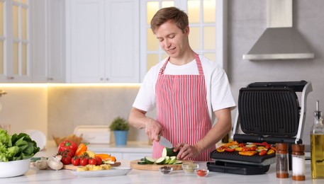 Photo of Man cutting zucchini while cooking vegetables on electric grill in kitchen