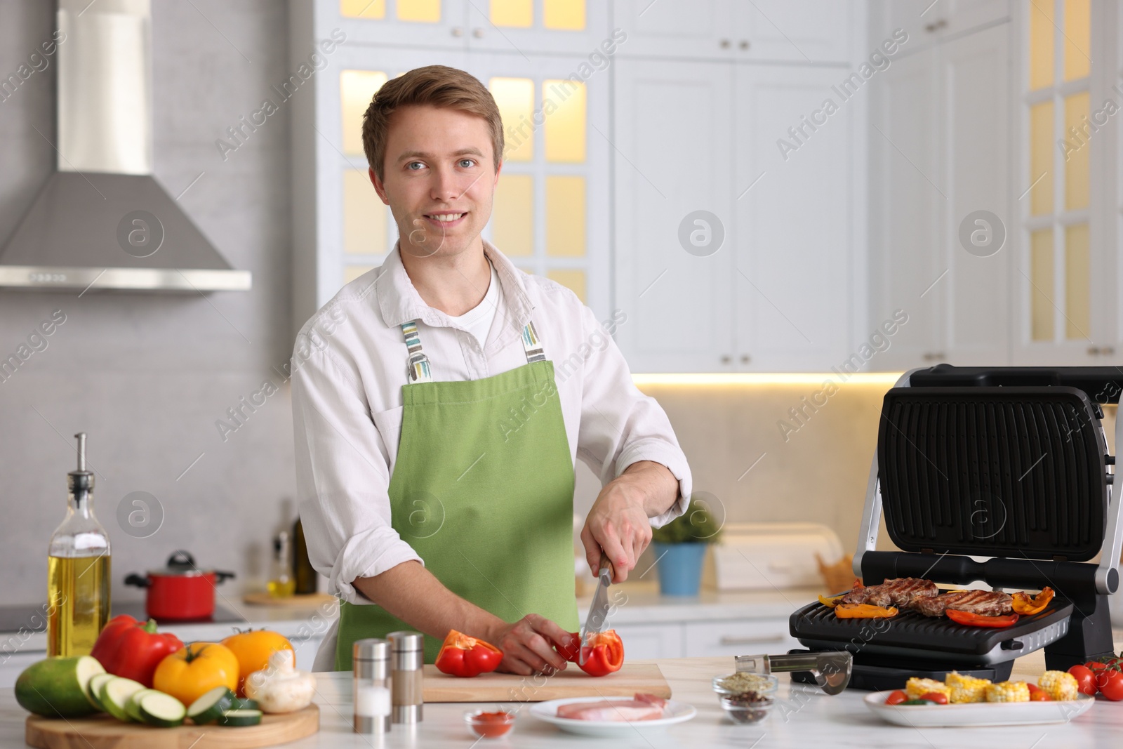 Photo of Man cutting pepper while cooking vegetables on electric grill in kitchen