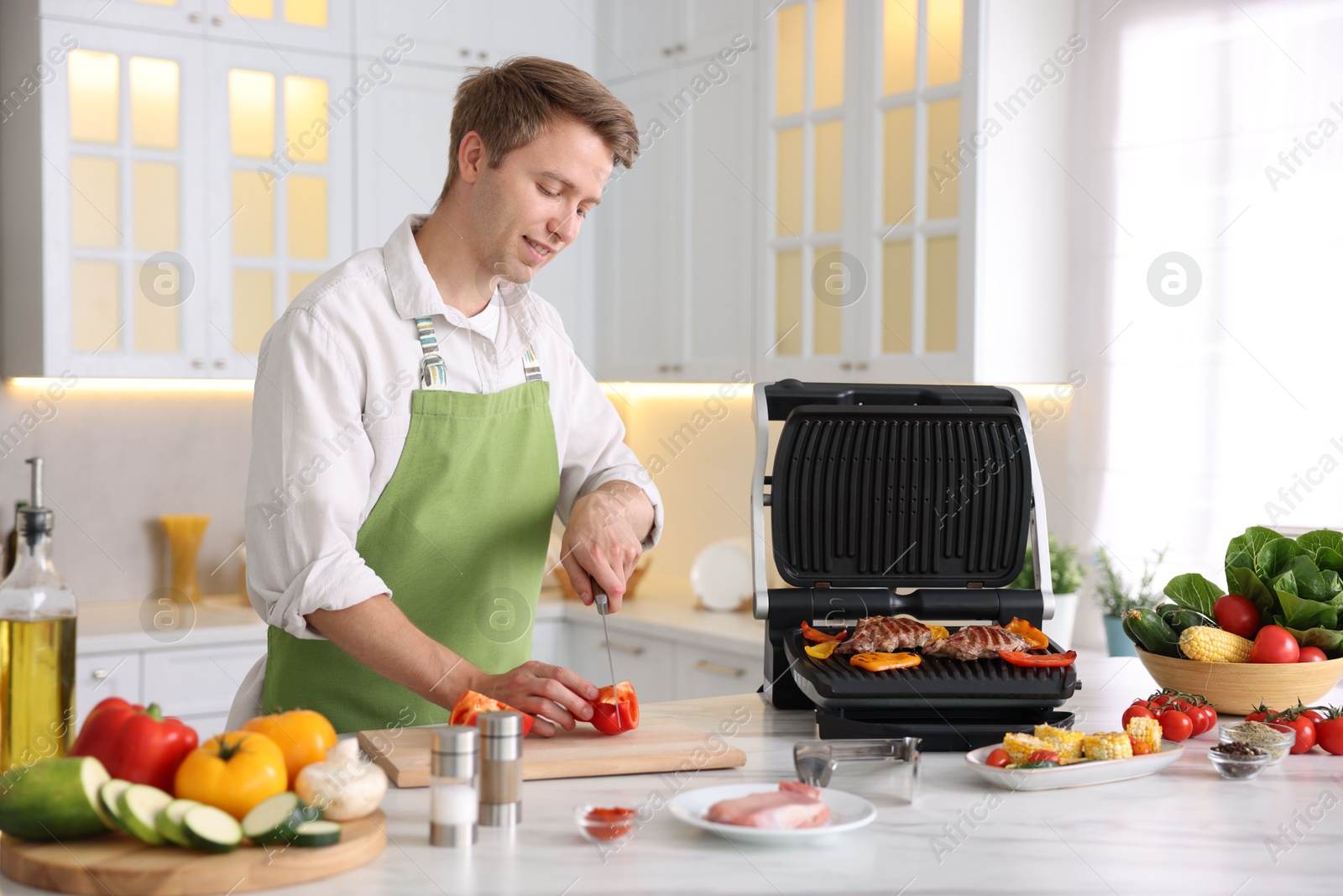 Photo of Man cutting pepper while cooking vegetables on electric grill in kitchen