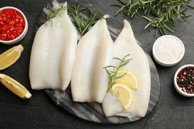 Photo of Uncooked squids with lemon slices, rosemary and spices on black table, flat lay