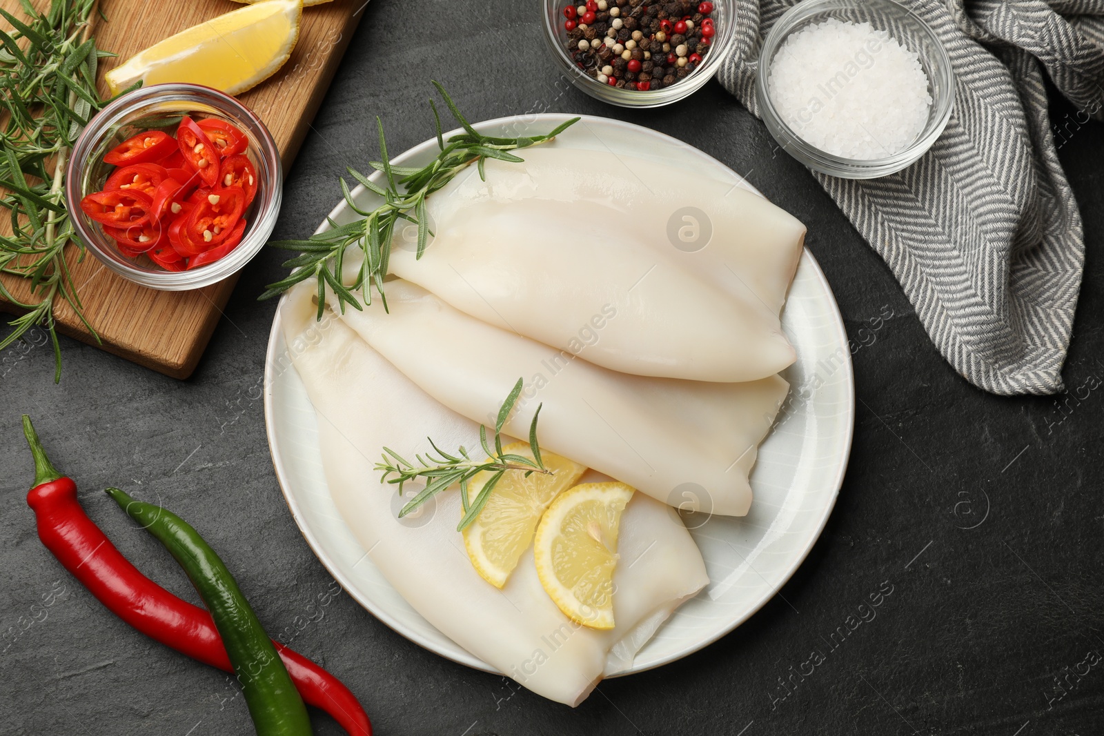Photo of Uncooked squids with lemon slices, rosemary and spices on black table, flat lay
